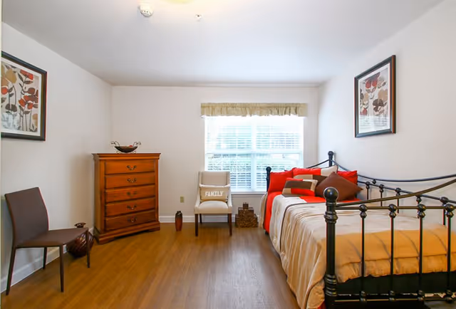 A bright bedroom with a black metal daybed adorned with red and brown pillows and a beige blanket. There is a wooden chest of drawers against the left wall, a beige chair with a 'FAMILY' pillow in front of a window with white blinds and a beige valance. Two framed floral artworks hang on the walls, and the floor is wooden.