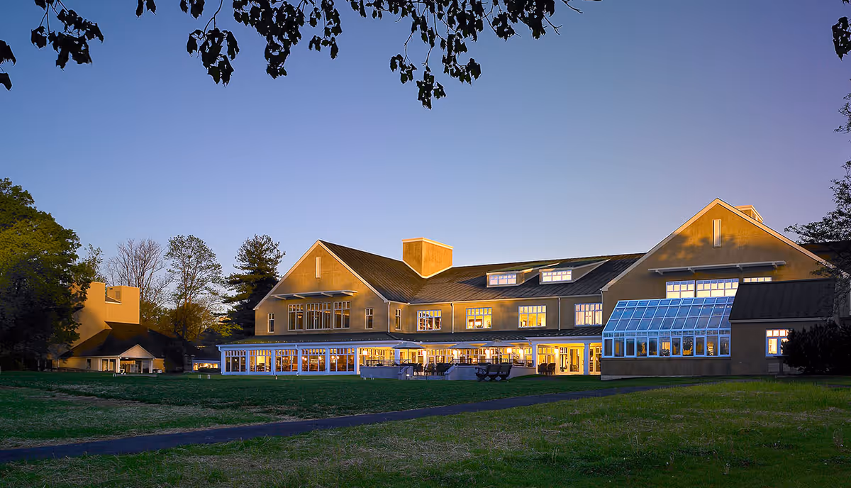 Exterior view of a large senior living facility building at dusk with lights glowing warmly from inside. The building has multiple windows, a glass sunroom, and a sloped roof. There is a grassy lawn and a paved pathway in the foreground, with tree branches hanging from the top of the image.