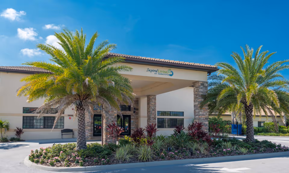 Exterior front view of Inspired Living Sarasota facility with a covered entrance supported by stone pillars, surrounded by palm trees and landscaped plants under a clear blue sky.
