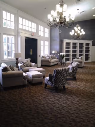 A spacious and well-lit living room area with large windows, multiple armchairs and sofas arranged around small tables, two chandeliers hanging from the ceiling, and a large white bookshelf against the far wall.