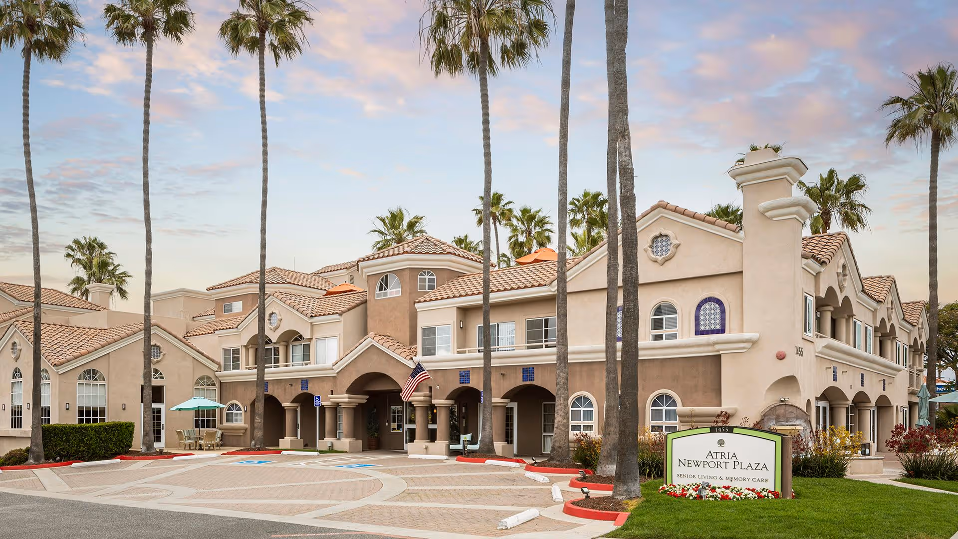Exterior view of Atria Newport Plaza, a senior living and memory care facility, featuring a large two-story building with beige and brown stucco walls, arched windows, and a tiled roof. Tall palm trees surround the building, and there is a paved driveway and parking area in front with a sign displaying the facility's name.