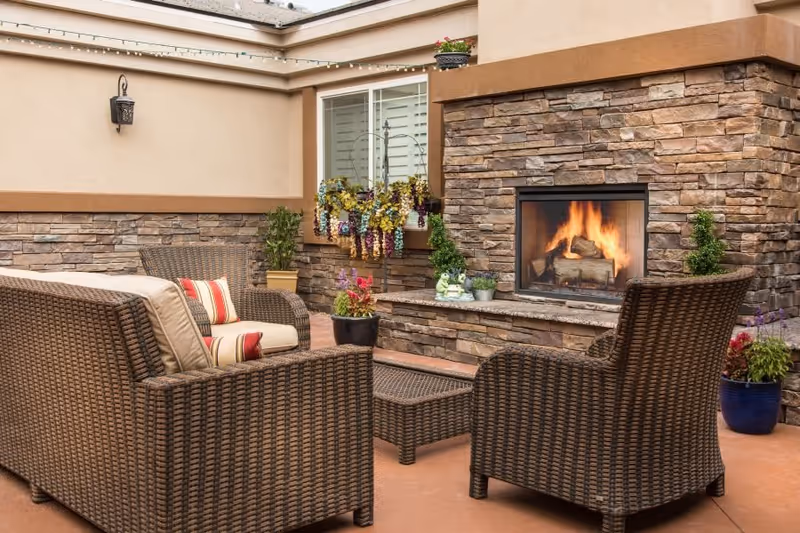 Outdoor patio area with a stone fireplace burning firewood, surrounded by wicker furniture including a sofa and two chairs with cushions and pillows. There are potted plants and hanging flowers near a window with white shutters on the beige wall.