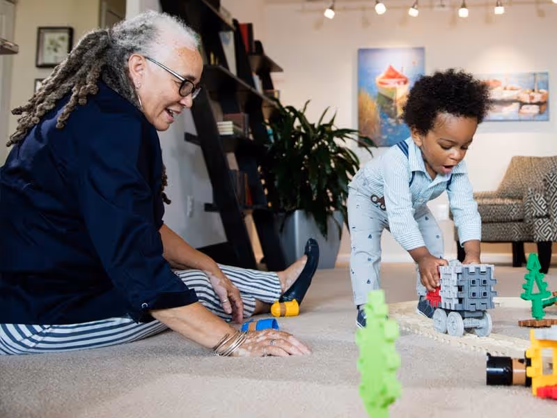 An elderly woman with gray dreadlocks and glasses sits on the carpeted floor playing with a young child who is pushing a toy train in a cozy living room setting with plants, artwork, and a patterned armchair in the background.