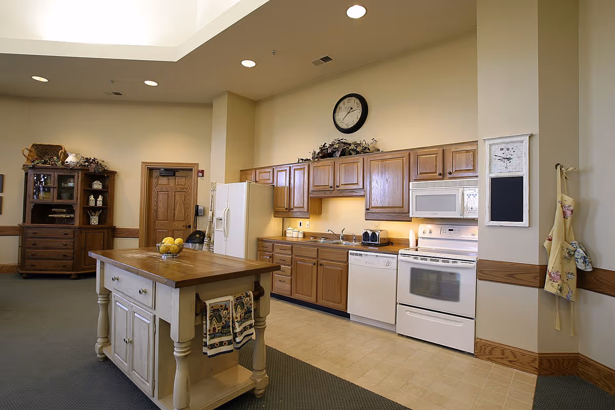 A spacious kitchen area in a senior living facility featuring wooden cabinets, a white refrigerator, dishwasher, stove, and microwave. There is a wooden island in the center with a bowl of lemons on top and decorative towels hanging on the side. The walls are light-colored with a clock above the cabinets and aprons hanging on the right wall. A wooden hutch with decorative items is visible in the background.