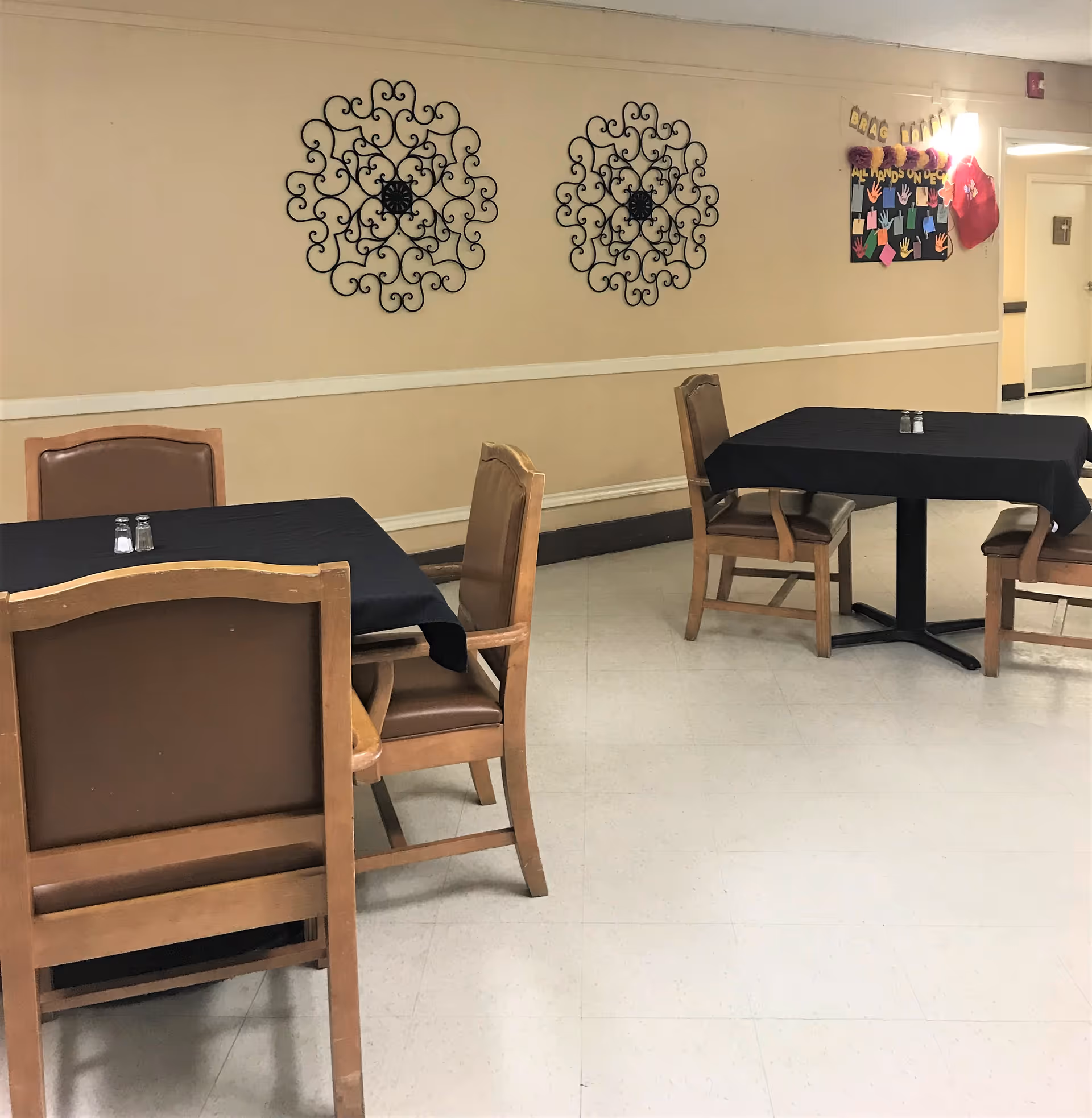 Interior view of a dining area with two tables covered with black tablecloths, each surrounded by four wooden chairs with brown cushions. The walls are beige with two decorative black wrought iron wall hangings and a bulletin board with colorful paper hands and letters. The floor is light-colored tile.
