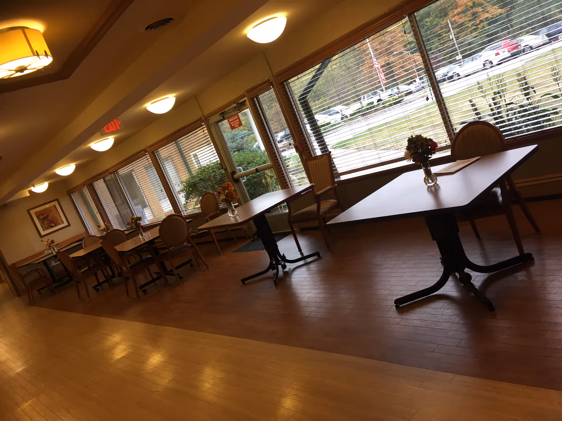 Interior view of a dining area with several tables and chairs arranged along large windows with blinds. Each table has a small vase with flowers. The room has wooden flooring and ceiling lights, and an exit sign is visible above a door.