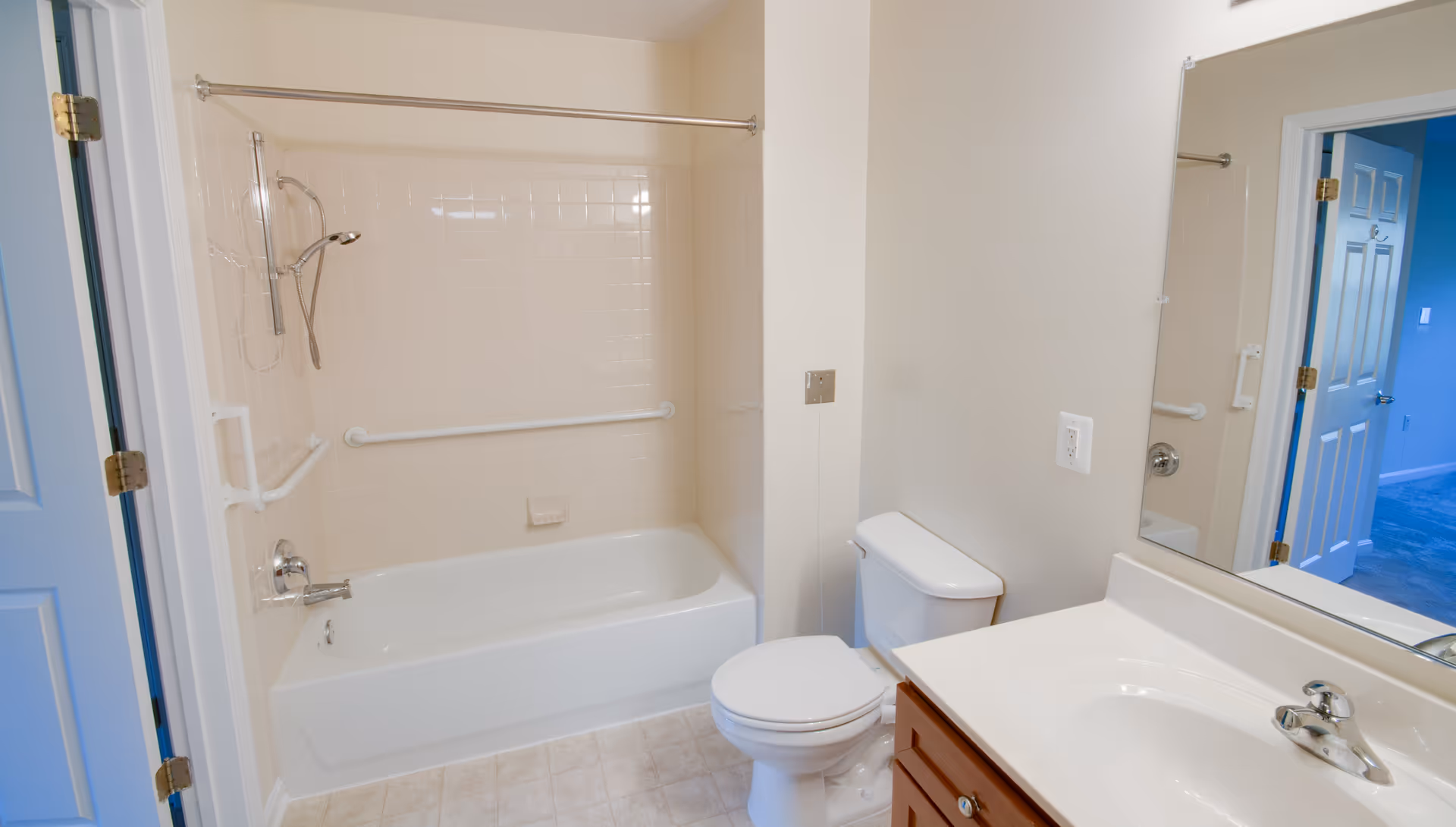 A clean bathroom featuring a white bathtub with a showerhead and grab bars, a white toilet, and a vanity with a sink and faucet. The walls are light-colored, and there is a large mirror above the sink. The bathroom door is open, showing a glimpse of a blue room outside.