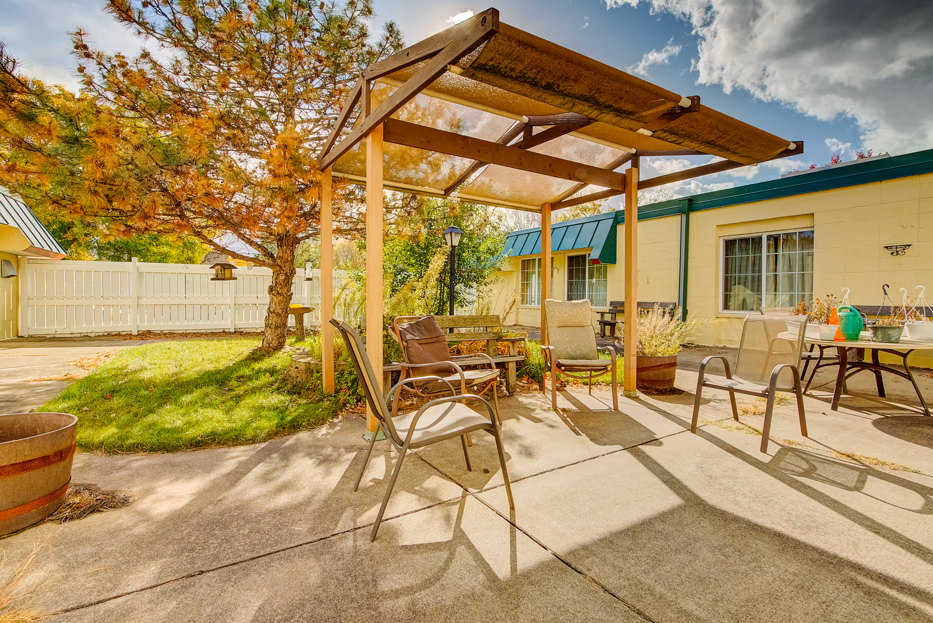 Outdoor patio area with several chairs arranged under a wooden pergola. The patio is surrounded by a grassy area with trees and a white fence. The building in the background has yellow walls and green awnings over the windows. There are also some potted plants and a table with gardening supplies on the right side.