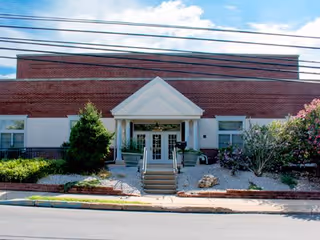 Front exterior view of a brick building with a white triangular pediment above the entrance, steps leading up to double glass doors, and landscaping with bushes and flowers on either side.