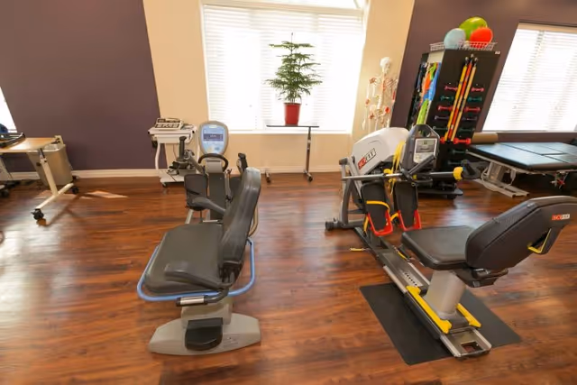 A rehabilitation exercise room with two recumbent exercise bikes, a treatment table, a skeleton model, and a rack holding colorful exercise bands and balls. The room has wooden flooring and large windows with blinds letting in natural light.