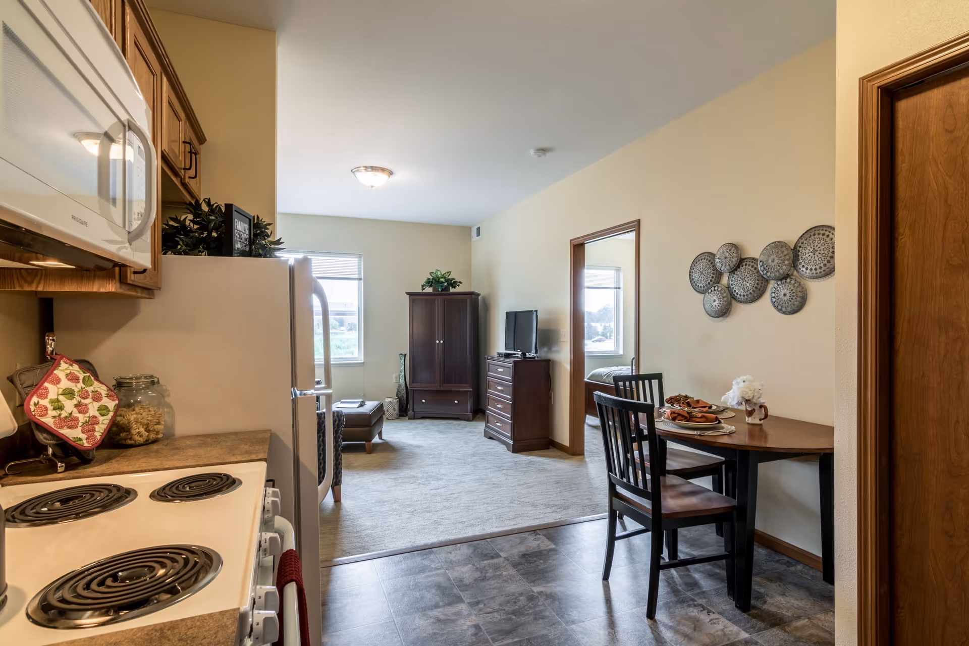 View of a senior living apartment interior showing a kitchen area with stove, microwave, and refrigerator on the left, a small dining table with two chairs on the right, and a living room area with a TV, cabinet, and window in the background. A bedroom is visible through an open doorway.