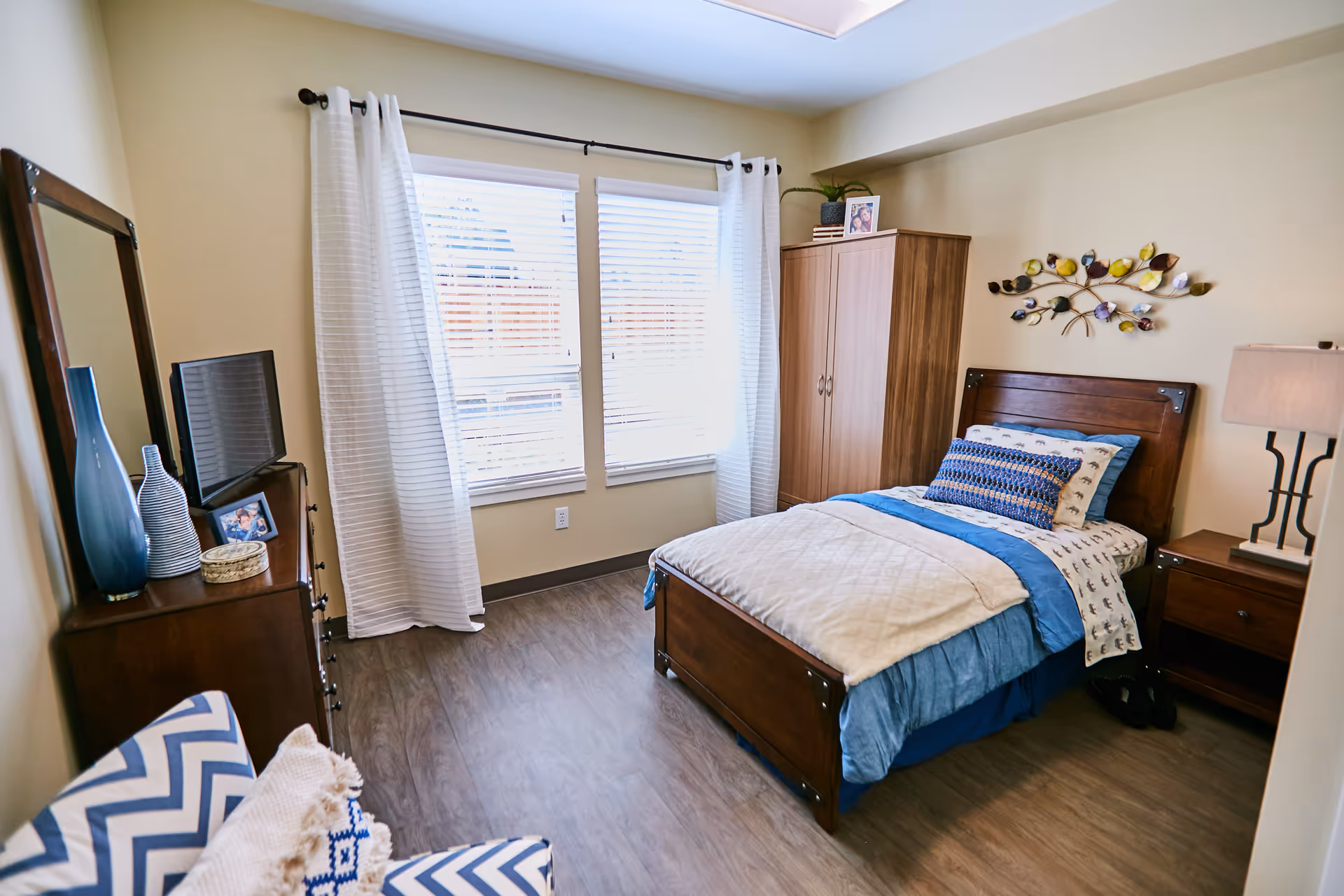 A cozy bedroom with a single wooden bed dressed in blue and white bedding, a wooden wardrobe, a nightstand with a lamp, a dresser with a TV and decorative vases, a large mirror, and a blue and white chevron-patterned chair. The room has light-colored walls, wood flooring, and two windows with white curtains letting in natural light.