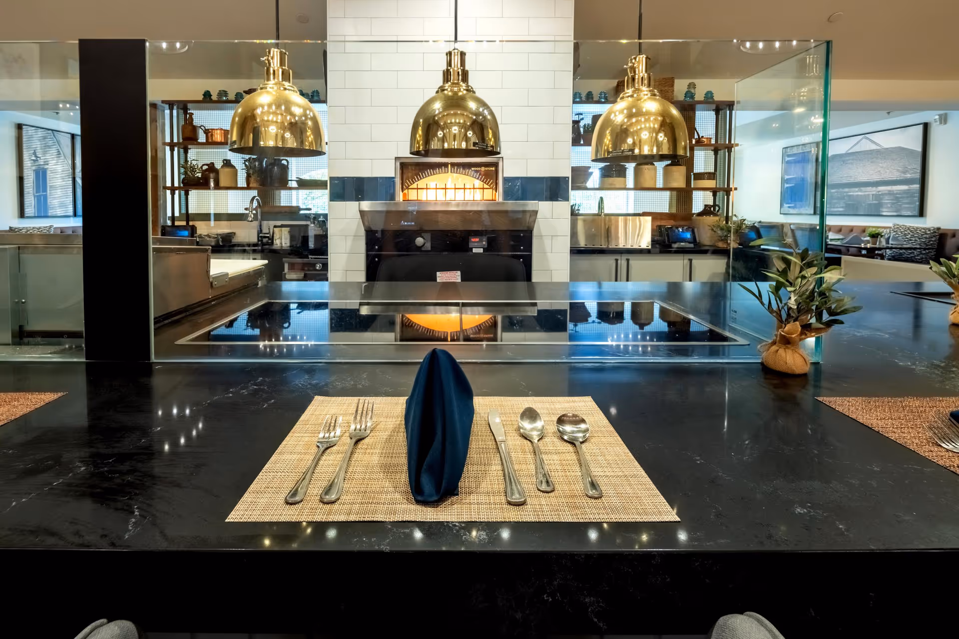 A single place setting on a black countertop facing an open kitchen with brass pendant lights and a tiled oven.