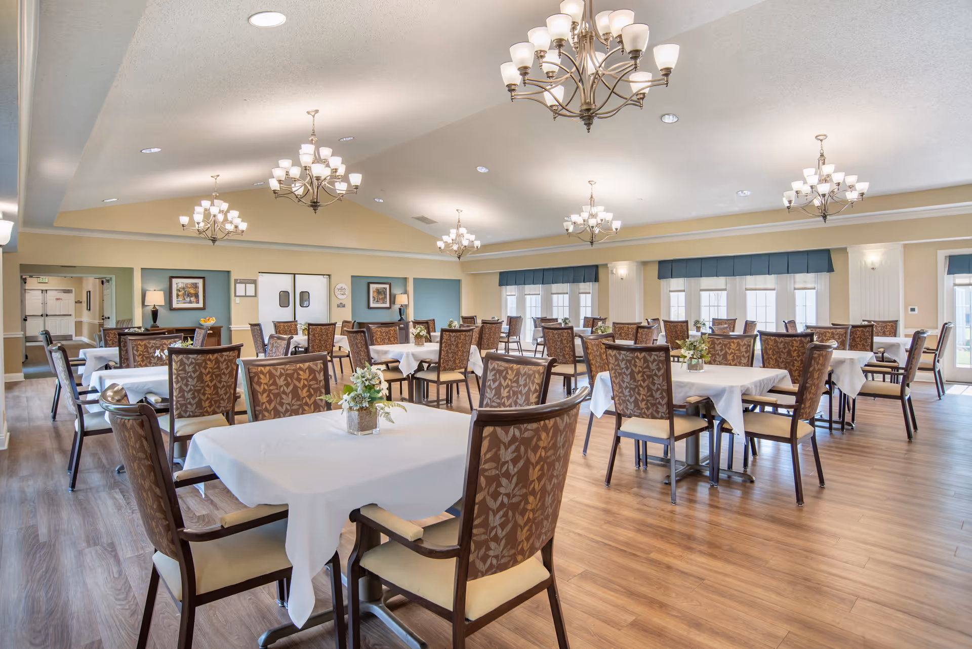 A spacious dining room with multiple tables covered in white tablecloths, each surrounded by patterned chairs. The room features wooden flooring, large windows with blue valances, and several elegant chandeliers hanging from the ceiling. Small floral centerpieces are placed on each table.