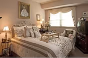 A cozy bedroom with a neatly made bed featuring multiple pillows and a beige and white floral bedspread. There is a wooden tray with a teapot and cups on the bed. To the right of the bed is a cushioned armchair with a pillow, and next to it is a dark wooden TV stand with an older-style television. A large window with beige curtains lets in natural light. The walls are decorated with framed pictures, including a portrait above the bed. A small side table with a lamp and decorative items is on the left side of the bed.