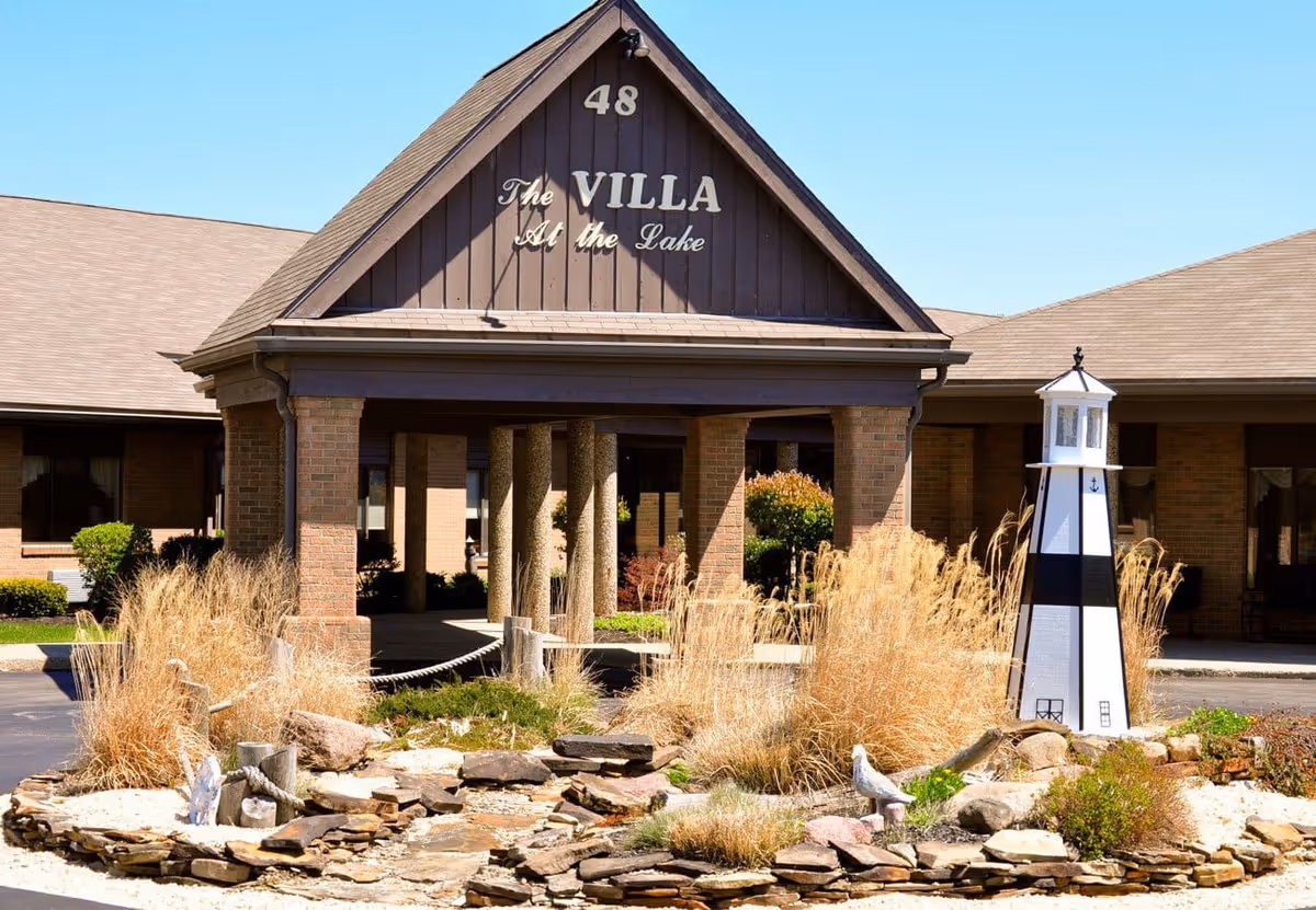 Exterior view of a senior living facility named The Villa At the Lake, showing the entrance with a peaked roof, brick columns, and a decorative lighthouse surrounded by ornamental grasses and rocks.