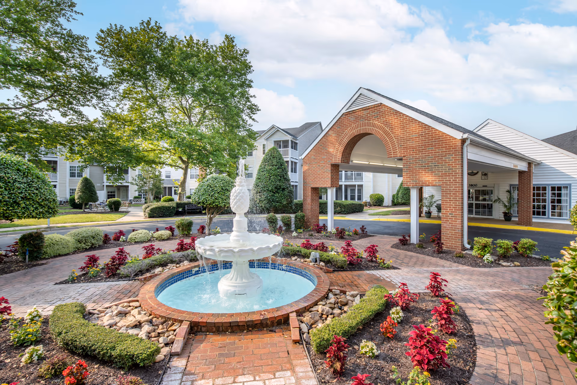 Front entrance of the senior living community featuring a brick porte-cochère, a central fountain, and landscaped flower beds.