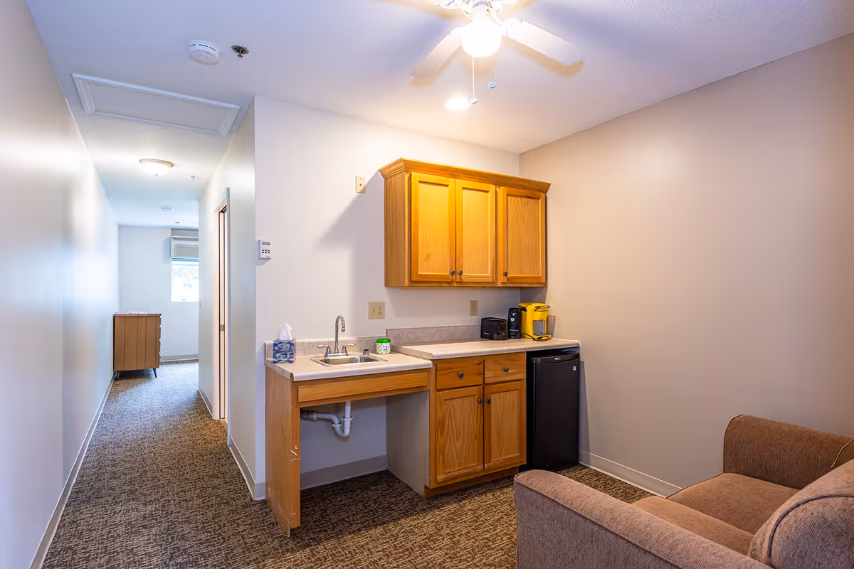 Interior view of a small kitchenette area in an assisted living facility with wooden cabinets, a countertop with a sink, a coffee maker, a mini refrigerator, and a brown upholstered chair. A hallway with carpet leads to another room with a window and a wooden dresser.