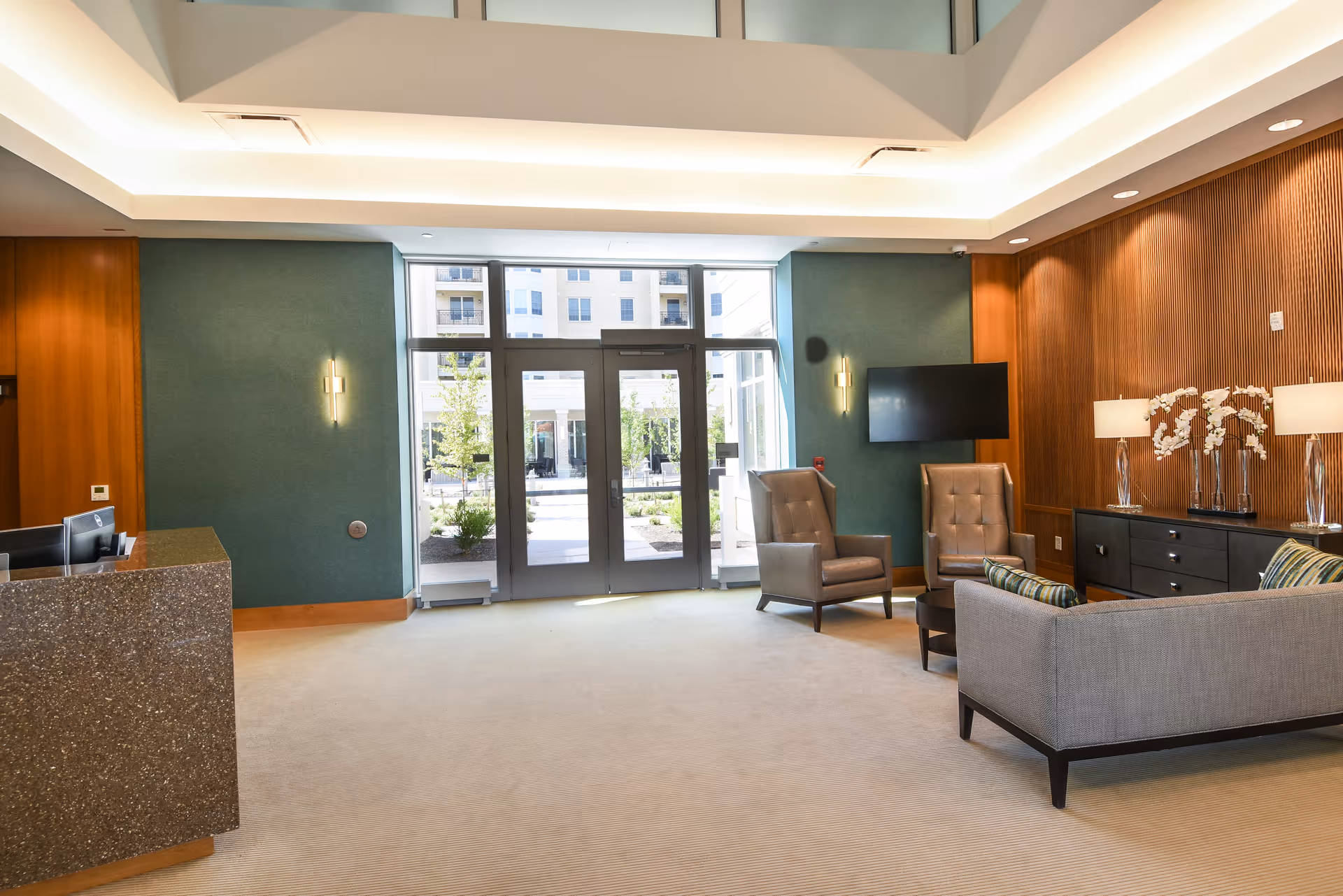 A modern and spacious lobby area with a reception desk on the left, large glass doors leading outside, and a seating area on the right featuring two armchairs, a sofa, a coffee table, and a sideboard with lamps and decorative flowers. The walls are a mix of green and wood paneling, and there is a flat-screen TV mounted on the wall.