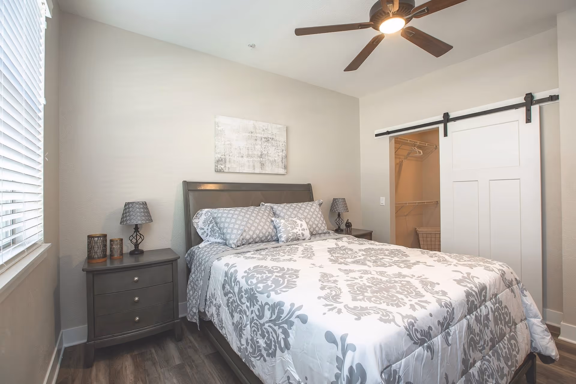 A neatly made bed with a white and gray patterned comforter and pillows in a bedroom. There are two matching nightstands on either side of the bed, each with a lamp. A window with white blinds is on the left wall, and a ceiling fan with a light is mounted on the ceiling. A sliding barn door reveals a walk-in closet with wire shelving and hangers.