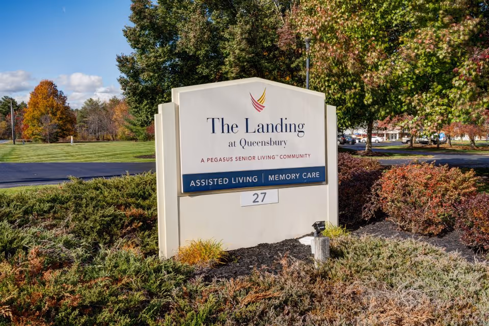 Outdoor view of a large sign for The Landing at Queensbury, a Pegasus Senior Living community offering assisted living and memory care. The sign is surrounded by bushes and trees with autumn foliage, and a parking lot is visible in the background.