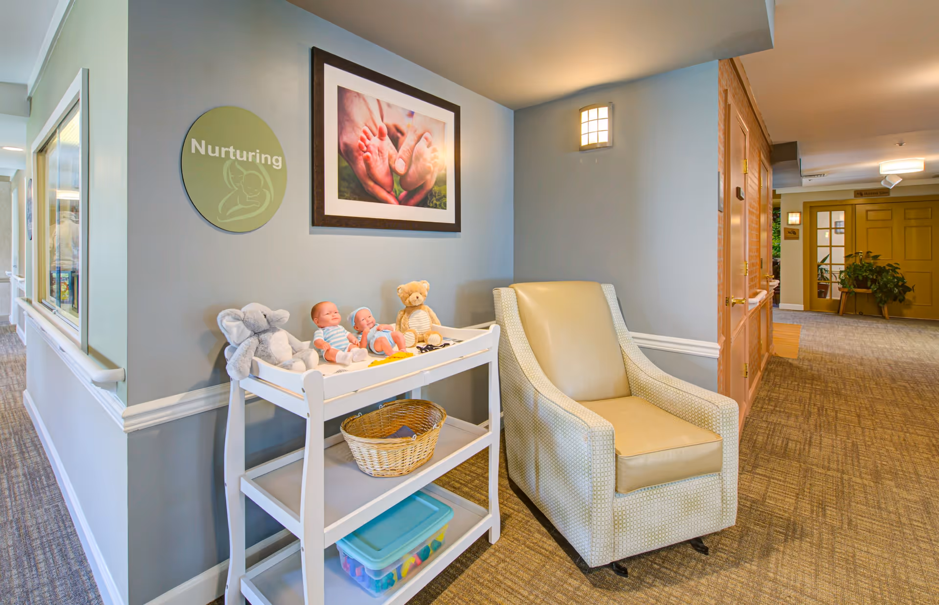 A cozy corner in a senior living facility with a beige armchair next to a white changing table holding baby dolls, stuffed animals, and a basket. The wall behind features a round sign that says 'Nurturing' and a framed photo of adult hands holding baby feet. The hallway extends to the right with doors and plants visible.