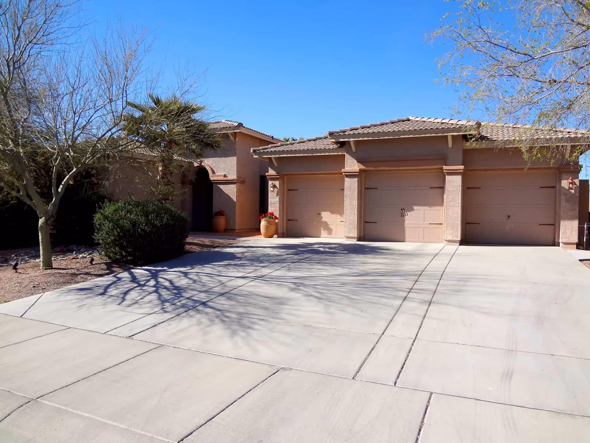 Exterior view of a single-story building with a three-car garage, beige stucco walls, and a tiled roof. There are two large potted plants with red flowers near the entrance, and leafless trees and bushes surround the driveway under a clear blue sky.