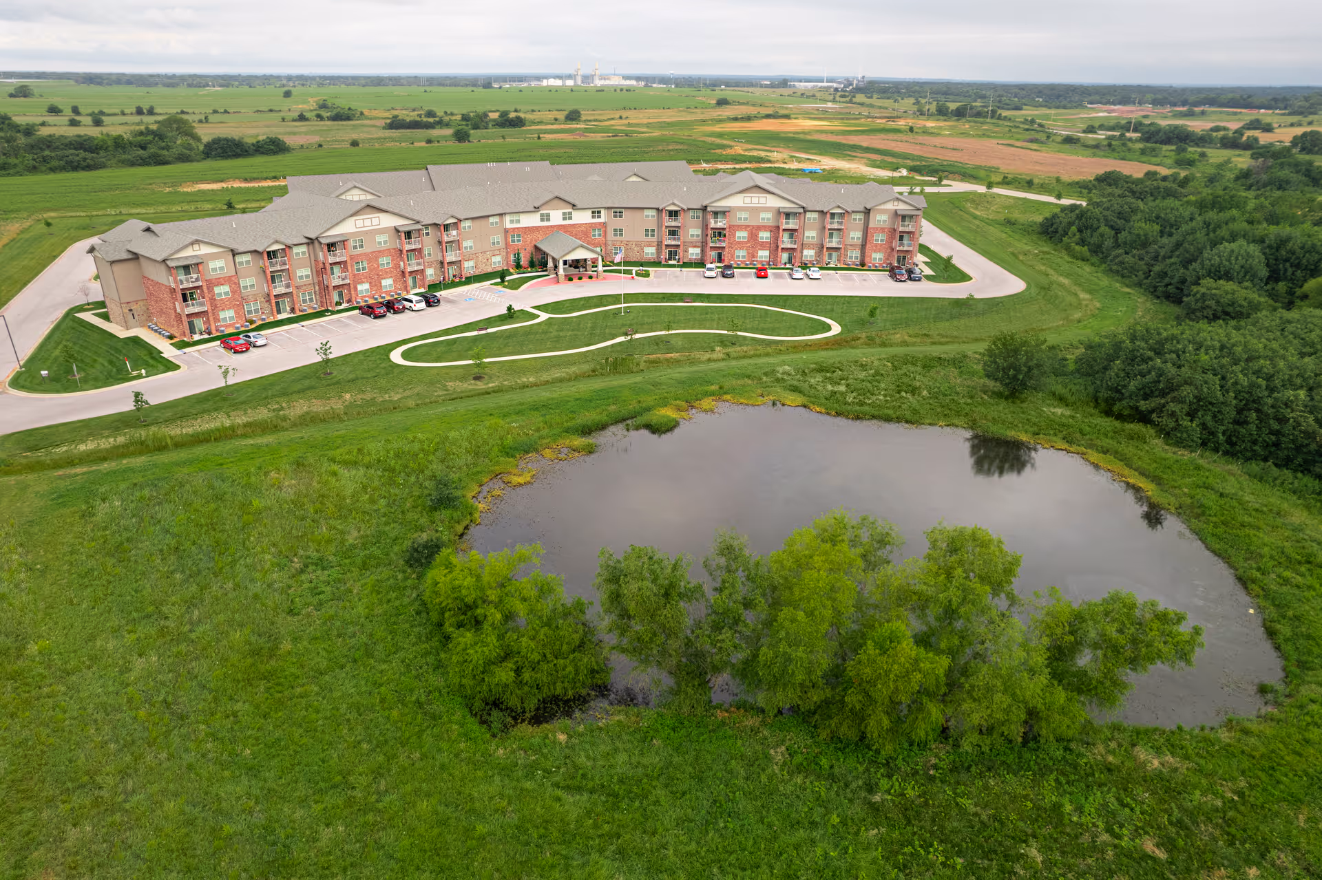 Aerial view of The Wildwood Senior Living facility showing a large three-story building with a gray roof and brick exterior surrounded by green lawns, a small pond with trees, a curved driveway, and several parked cars. The background features expansive farmland and a cloudy sky.