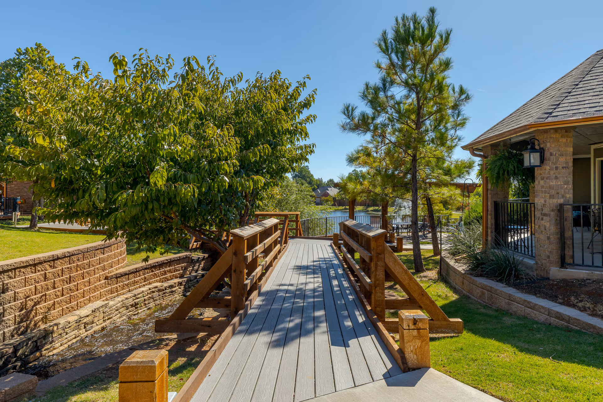 A wooden footbridge over a small stream in a landscaped outdoor area with green grass, trees, and a brick building with a covered porch on the right. In the background, there is a lake with a fountain and outdoor seating under a pergola.