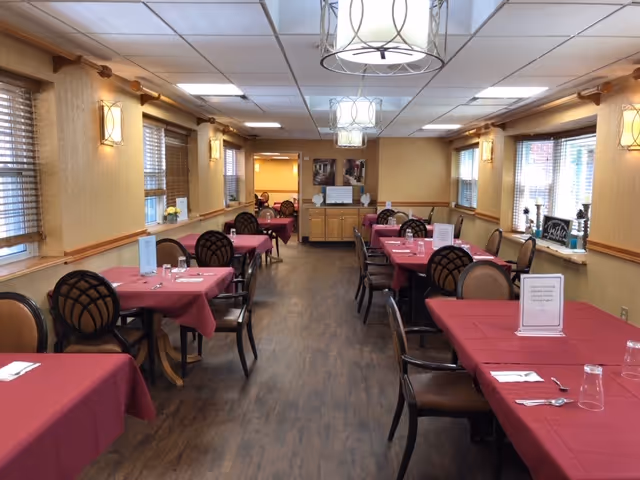 Dining room with multiple tables covered in red tablecloths, each set with utensils, glasses, and menus. The room has wooden floors, beige walls with windows on both sides, and ceiling lights with decorative fixtures. Chairs with cushioned seats and backs are arranged around the tables.