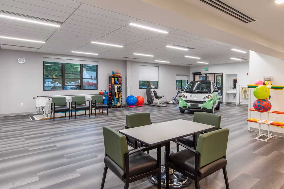 A spacious rehabilitation room with exercise equipment including stationary bikes, therapy balls, and a small green and white car. There are four green and black chairs around a table in the foreground, and additional chairs lined up against the wall. The room has large windows with a view of trees and houses outside, and the floor is covered with gray wood-like vinyl.