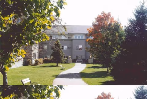 A paved walkway leads through a grassy courtyard area with benches and trees, including one with autumn-colored leaves, towards a multi-story residential building under a clear sky.