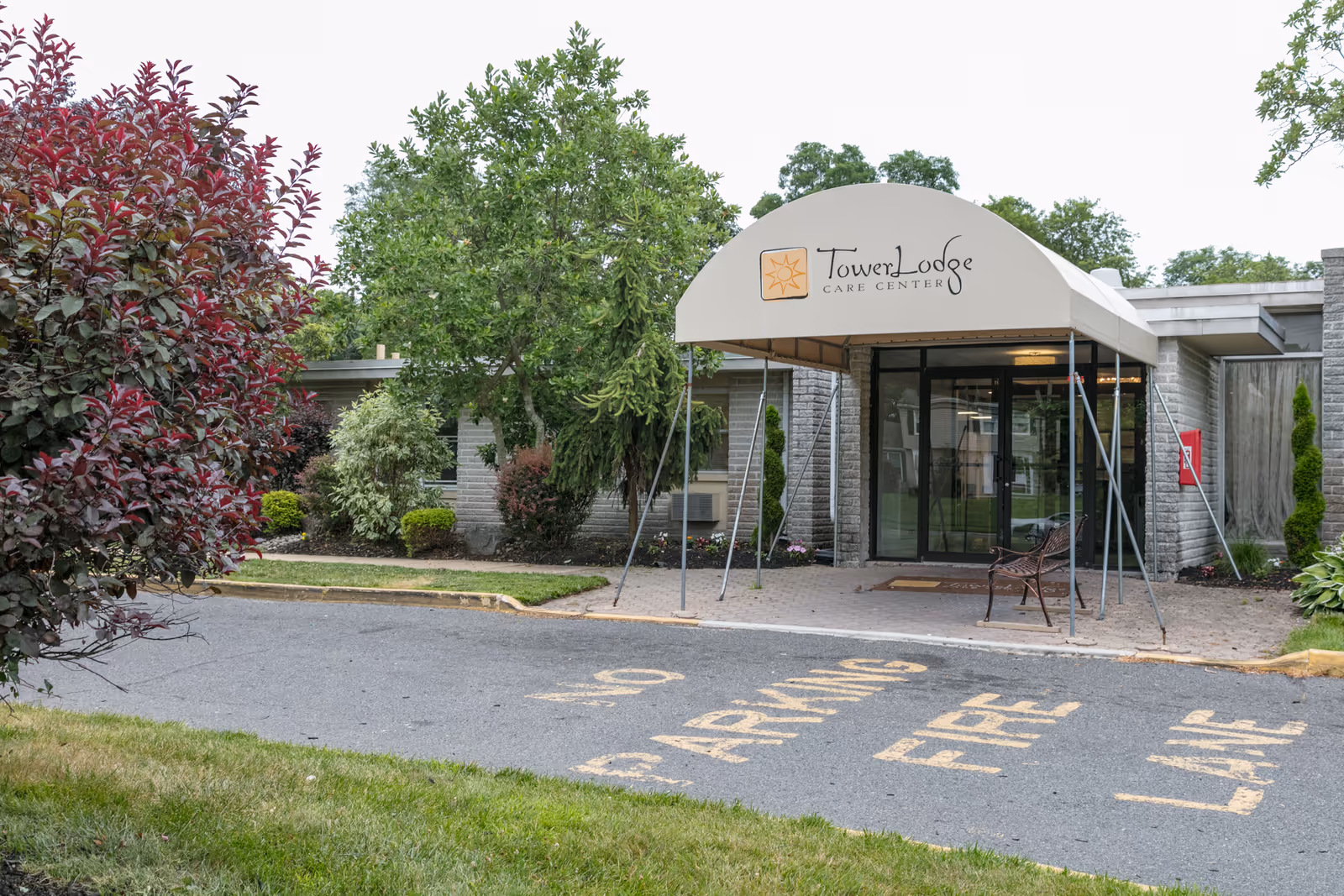 Entrance of Tower Lodge Care Center with a beige canopy displaying the facility's name and logo. The entrance has glass doors and is surrounded by greenery including trees and bushes. There is a bench near the entrance and a paved driveway with yellow painted words indicating no parking and fire lane.