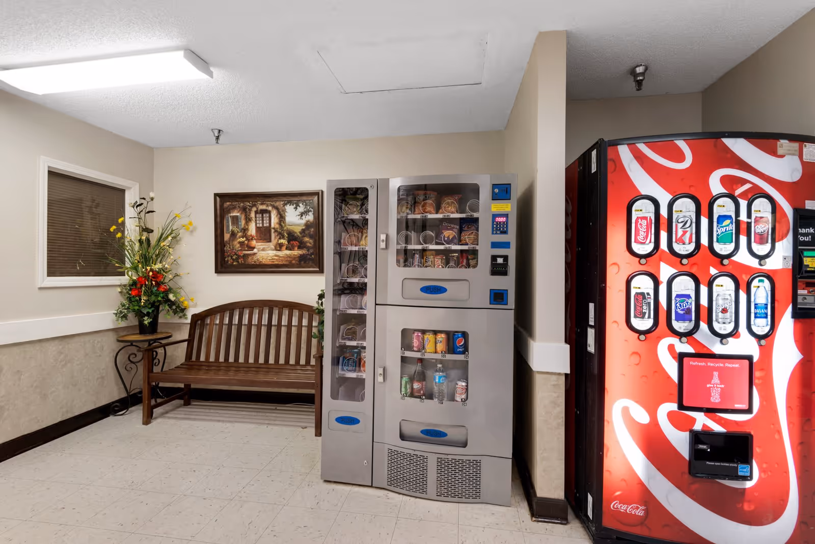 Indoor corner area with a wooden bench, a side table with a flower arrangement, a framed painting on the wall, a snack vending machine, and a Coca-Cola beverage vending machine.