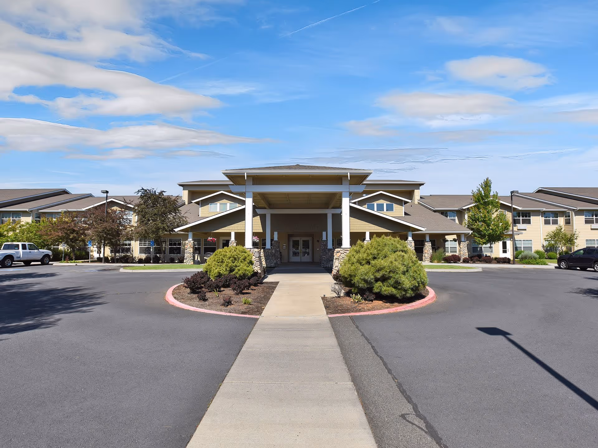 Front exterior view of Prestige Senior Living High Desert building with a covered entrance, landscaped bushes, and a clear blue sky with some clouds.