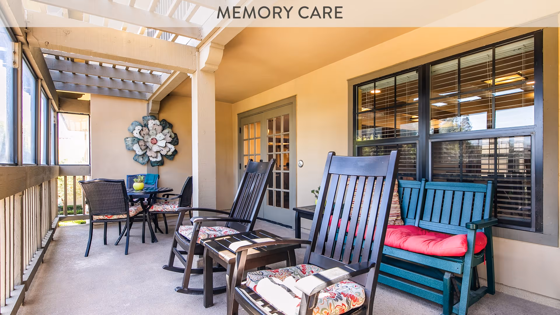 Covered outdoor patio area with rocking chairs, a small table with chairs, and a decorative wall flower. The space is well-lit with natural light coming through the windows and has a beige wall with a door leading inside.