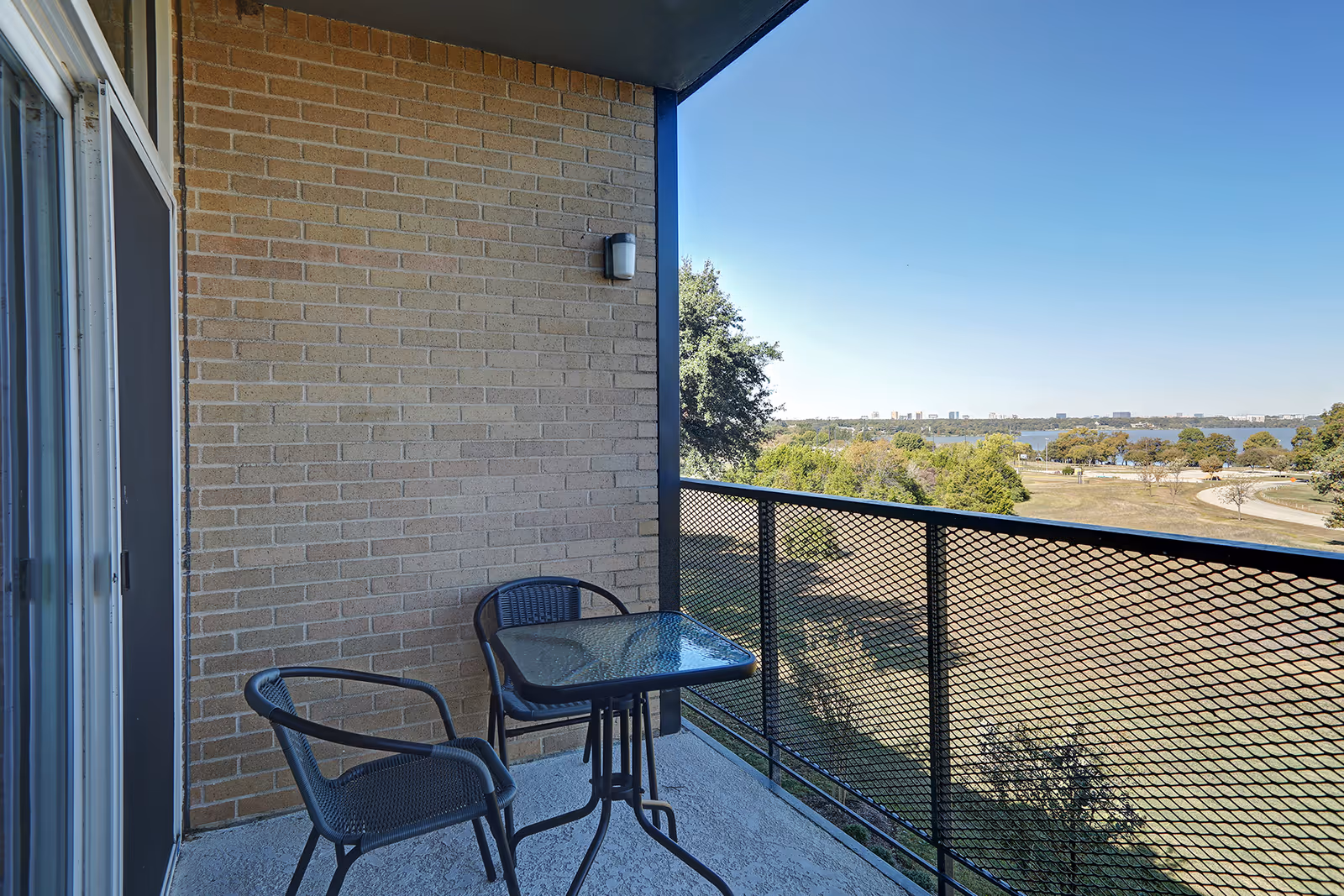 A small outdoor balcony with a glass-top table and two black chairs. The balcony has a black metal mesh railing and overlooks a park area with trees and a body of water in the distance under a clear blue sky.