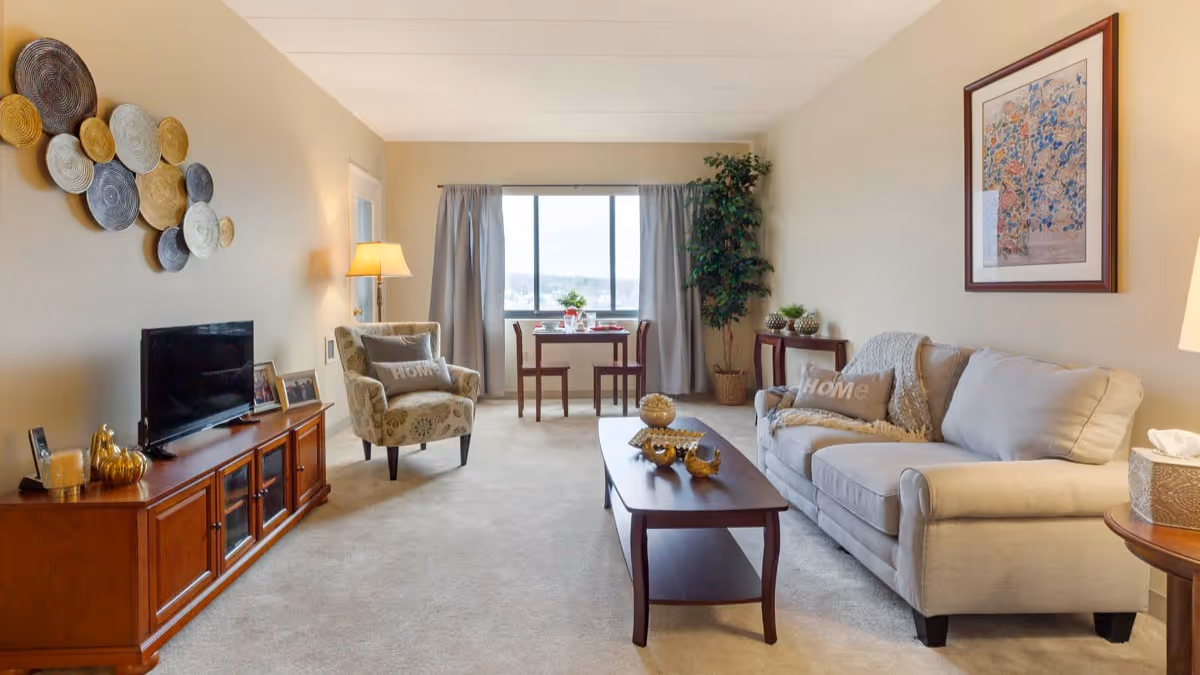 A cozy living room in a senior living facility featuring a beige sofa with a 'Home' pillow, a patterned armchair, a wooden coffee table with decorative items, a wooden TV stand with a flat-screen TV and framed photos, a floor lamp, a small dining table with two chairs near a window with curtains, a potted plant, and wall art including a framed picture and decorative circular wall hangings.