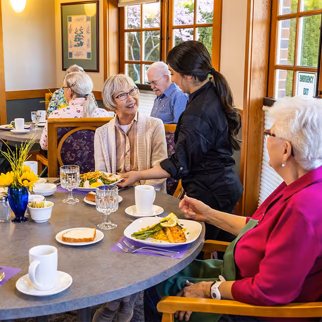 A dining room scene at The Terraces at Summitview where elderly residents are seated around a table enjoying a meal. A staff member is serving food to a smiling elderly woman. The table is set with plates of food, glasses, cups, and a small vase with yellow flowers. Large windows and framed artwork are visible in the background.