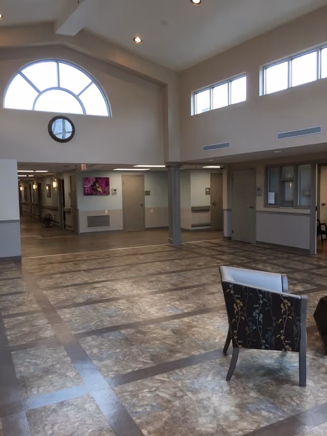 Interior view of a spacious common area in a senior living facility with high ceilings, large arched and rectangular windows allowing natural light, a clock on the wall, several closed doors, a hallway leading to other rooms, and a single upholstered chair with floral pattern in the foreground.
