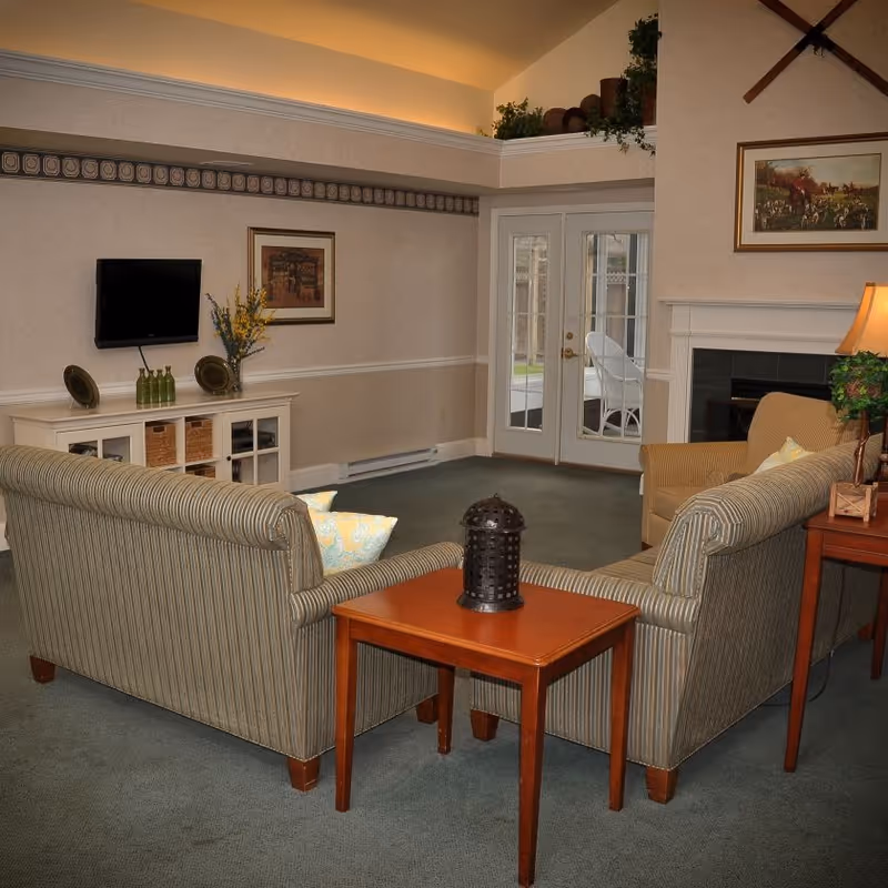A cozy living room with two striped sofas facing each other and a wooden side table between them. There is a fireplace on the right wall with a framed painting above it, a lamp on a side table, and French doors leading outside. On the left wall, a flat-screen TV is mounted above a white cabinet with decorative items and a vase with flowers.