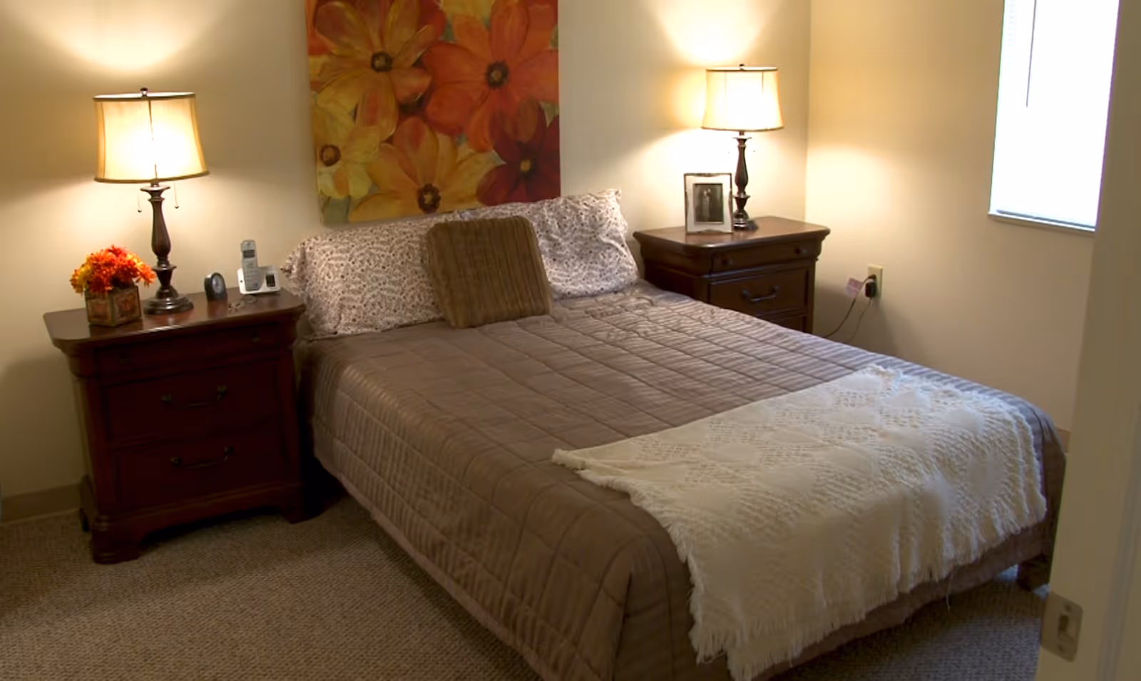 A neatly made bed in a warmly lit bedroom with two wooden nightstands, lamps, and floral artwork above the headboard.