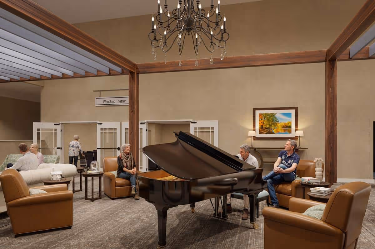 A spacious senior living facility common area with a grand piano in the center. Several elderly people are seated on comfortable leather chairs and sofas around the piano, engaging in conversation. The room features wooden beams, a chandelier, and a framed landscape painting on the wall. A sign above double doors reads 'Woodland Theater.'