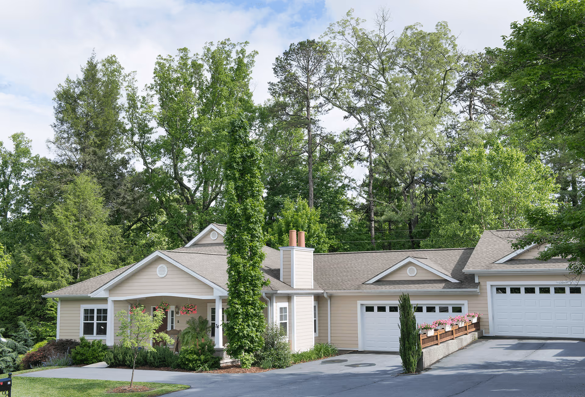 Exterior view of a single-story beige retirement community building with a front porch, surrounded by lush green trees and landscaping, under a partly cloudy sky.