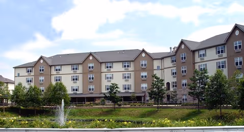 Exterior view of a large multi-story senior living facility building with beige and light brown siding, multiple windows, and a sloped roof. In the foreground, there is a landscaped area with green grass, trees, shrubs, and a small water fountain under a partly cloudy sky.
