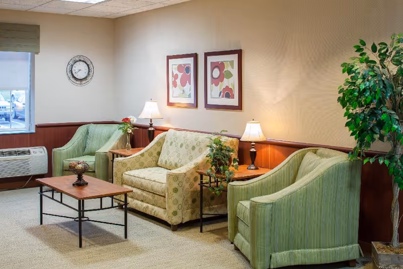 A cozy sitting area in a senior living facility with two green armchairs and a patterned loveseat arranged around a wooden coffee table. Two table lamps on side tables provide warm lighting. The walls are decorated with two framed floral prints, and a large potted plant is in the corner. A clock is mounted on the wall near a window with a view outside.