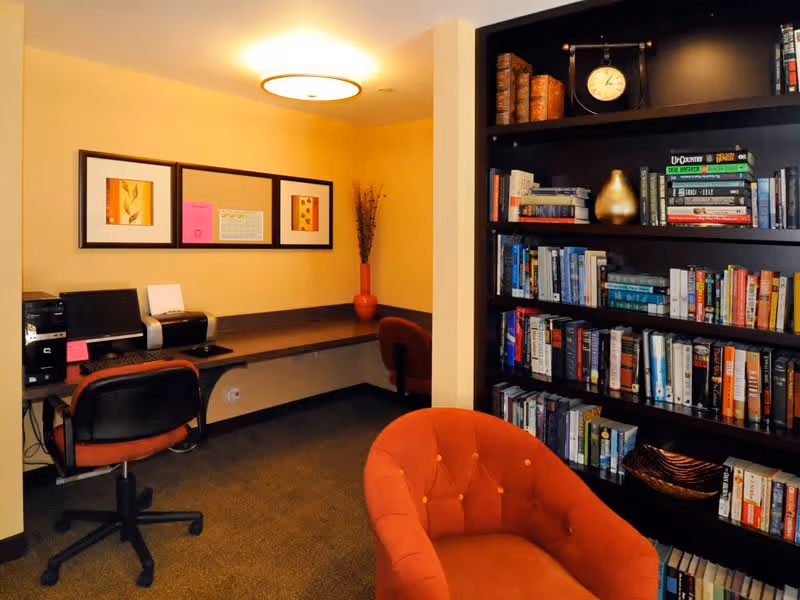 A cozy interior room featuring a computer workstation with a desktop computer, printer, and office chair against a beige wall with framed artwork. To the right, there is a large dark wooden bookshelf filled with books and decorative items, and in the foreground, an orange upholstered armchair.