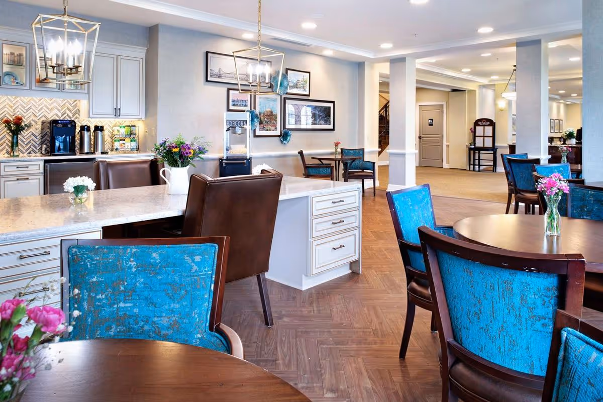 A bright and spacious dining area in a senior living facility featuring round wooden tables with blue upholstered chairs. There is a large kitchen island with brown leather chairs and a vase of flowers on top. The background shows a popcorn machine, framed artwork on the walls, and a hallway leading to other rooms.