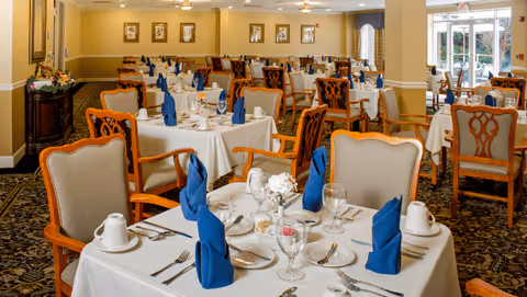 A spacious dining room with multiple tables covered in white tablecloths, each set with blue folded napkins, glassware, plates, and silverware. The room has beige walls with framed pictures and large windows letting in natural light. The chairs have wooden frames with beige upholstery.