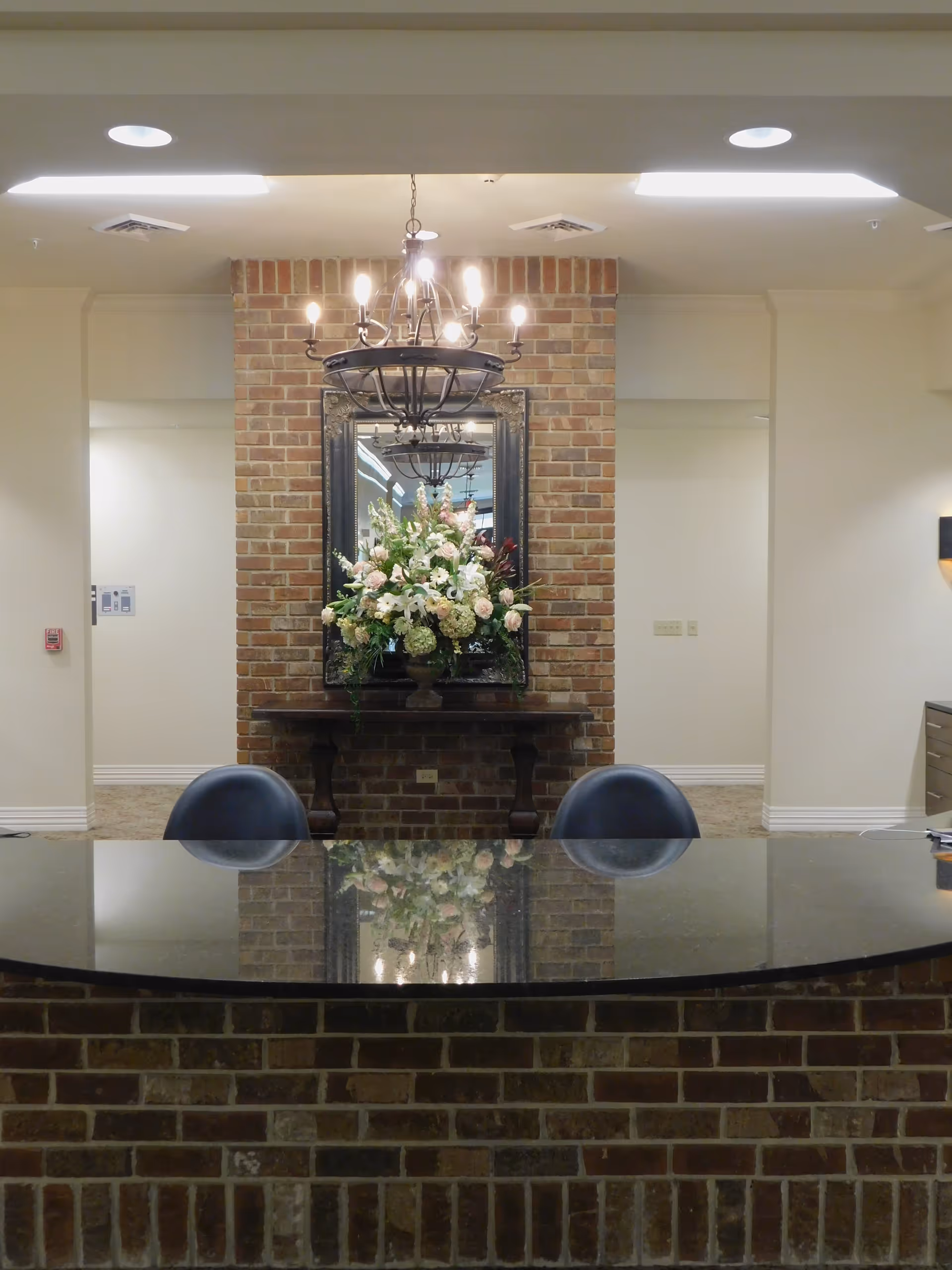 Reception area with a curved black countertop and two black chairs. Behind the counter is a brick wall with a large mirror and a floral arrangement on a wooden table. A chandelier with lit bulbs hangs from the ceiling above the flowers.